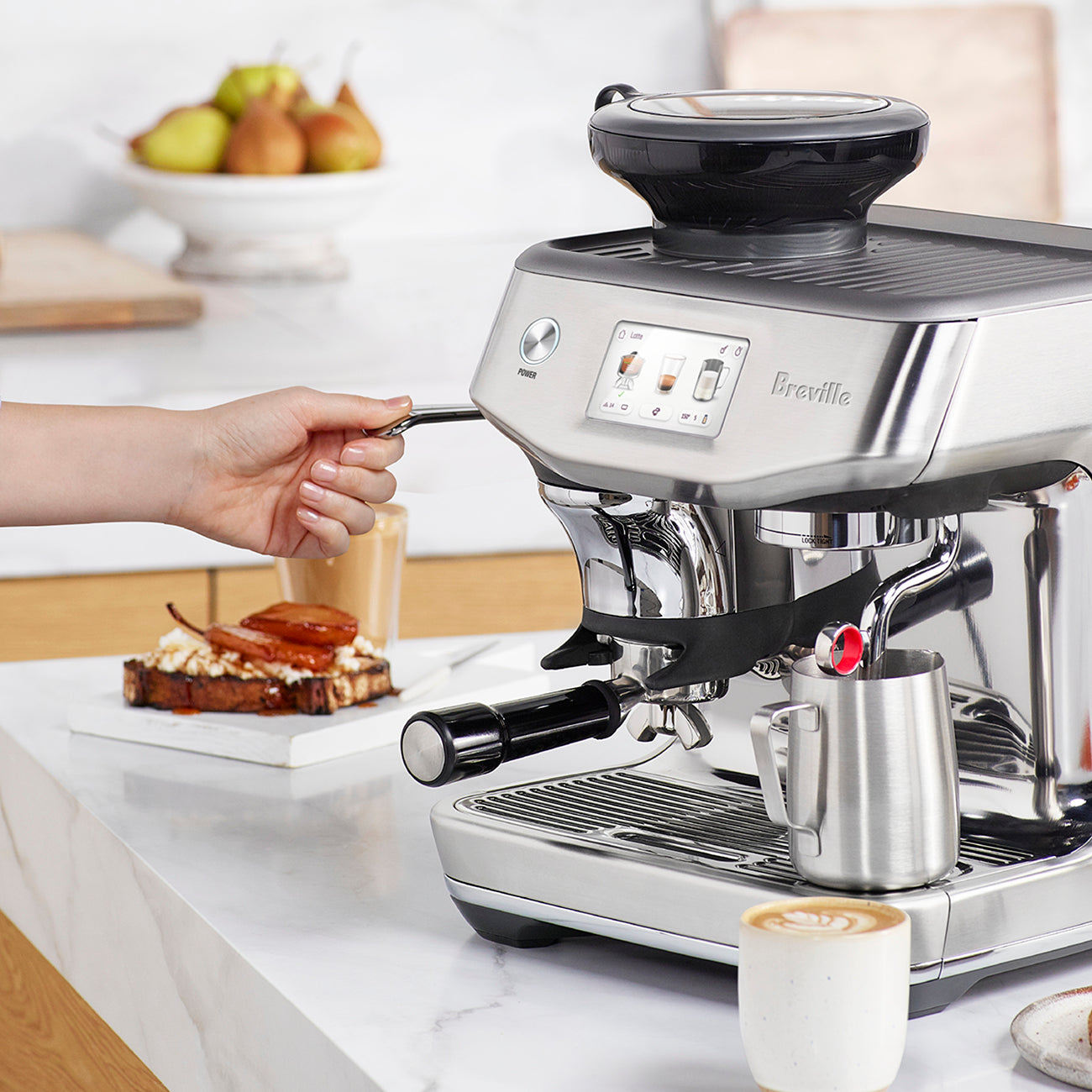 A person uses the steam wand of the Breville Barista Touch Impress Espresso Machine to make silky microfoam milk; a coffee cup, milk jug, and toast on the counter evoke a true morning barista ritual.