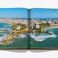 **Product Name: Biarritz Basque** by **Assouline** showcases an aerial photograph of Biarritz, a coastal city in the Basque Country. The image captures waves crashing against rocky shores, with buildings in the background—a paradise for surfing enthusiasts.