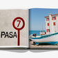 Open book showing two pages: the left page displays the text "PASA 7" with a red and black circular sign, while the right page features a white fishing boat near a seaside building with red shutters in the picturesque Basque Country. This image is from Biarritz Basque by Assouline.