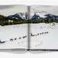An open book from Assouline titled "Big Sky" shows a snowy landscape with two dog sled teams crossing snow, framed by pine trees and mountains near Big Sky, Montana.