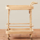 The Flore Bistro Bar Cart by Selamat, crafted from handwoven Manau rattan with two shelves, rounded edges, a curved handle, and four caster wheels, sits on a tile floor against a neutral wall.