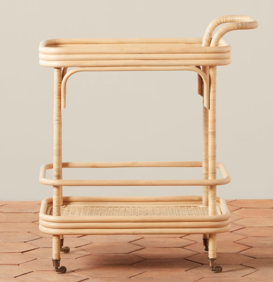 The Flore Bistro Bar Cart by Selamat, crafted from handwoven Manau rattan with two shelves, rounded edges, a curved handle, and four caster wheels, sits on a tile floor against a neutral wall.