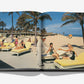 Open book from Assouline titled "The Boca Raton: A Century of Being Iconic," featuring a vintage photo of guests on yellow lounge chairs at the historic beachside resort, palms overhead and a waiter serving drinks.