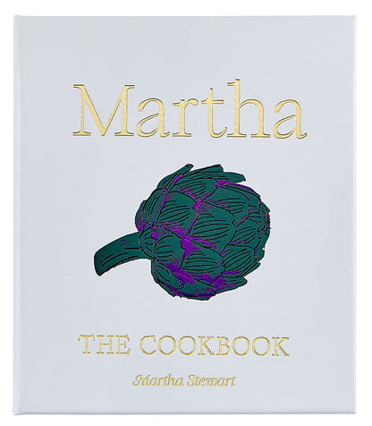 A white leather-bound cookbook by Graphic Image, titled "Martha: The Cookbook," features gold text and a central artichoke illustration, suggesting a curated collection of Martha Stewart’s favorite recipes.