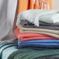 A stack of Pezzo Throw Collection blankets by Matouk—fringed and folded in light blue, gray, beige, coral, blue, and green soft brushed cotton—with more cozy throws draped in the background for a welcoming touch.