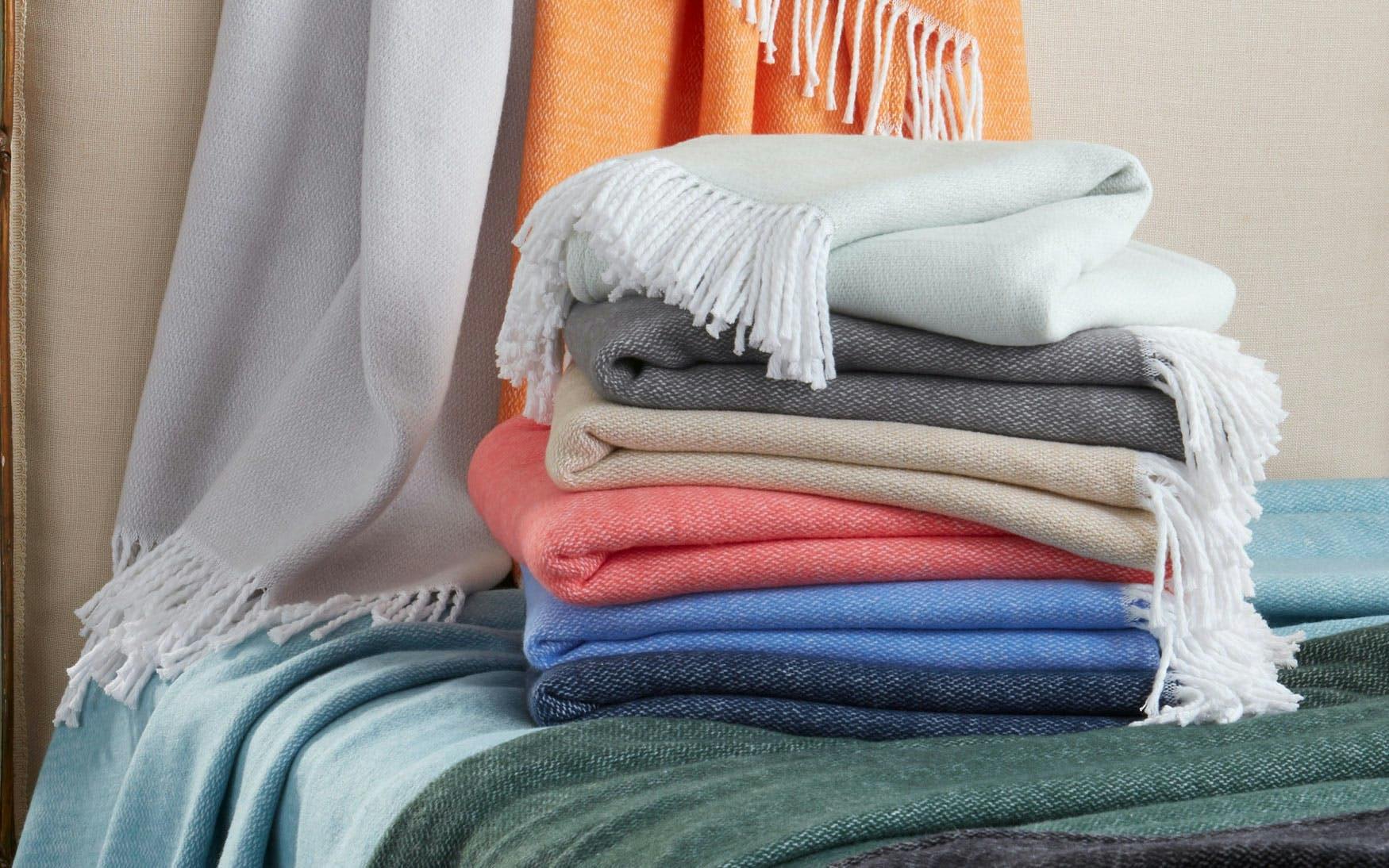 A stack of Pezzo Throw Collection blankets by Matouk—fringed and folded in light blue, gray, beige, coral, blue, and green soft brushed cotton—with more cozy throws draped in the background for a welcoming touch.