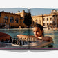 A person in a pool gazes intently at a chessboard on a floating platform, with the stunning Buda Castle visible in the background, embodying the allure of the Budapest Gem by Assouline.