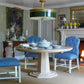A dining room with the Bunny Williams Haven Dining Table in cerused oak with a pedestal base, blue upholstered chairs, a statement green and gold chandelier, a fireplace, and wall art.