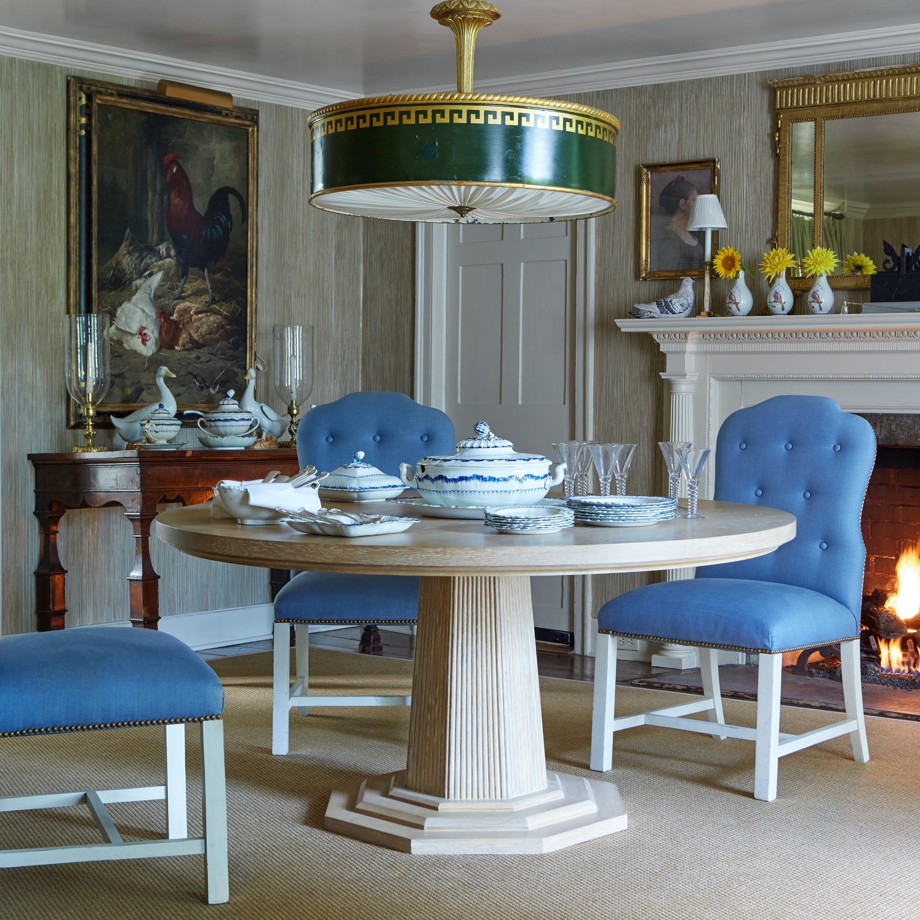 A dining room with the Bunny Williams Haven Dining Table in cerused oak with a pedestal base, blue upholstered chairs, a statement green and gold chandelier, a fireplace, and wall art.