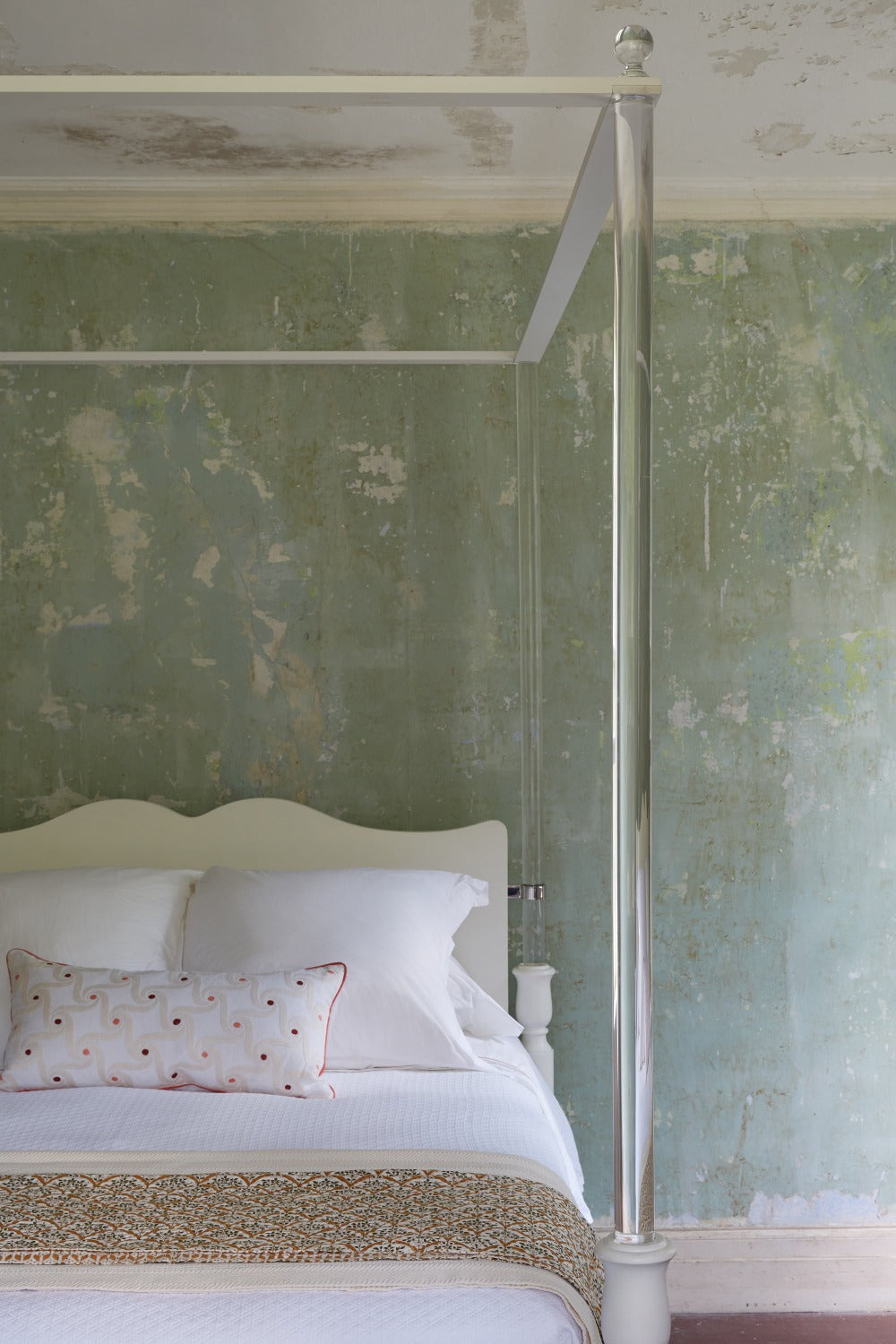The Garbo King Bed by Bunny Williams features cylindrical posts and spherical finials, shown here in white lucite against a distressed green textured wall, topped with white bedding and a decorative pillow.
