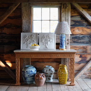 A wood table stands by a rustic wall, topped with a lamp, a bowl of lemons, and two Bunny Williams Pomegranate Plaster Panels; five painted ceramic vases are arranged on the floor beneath.