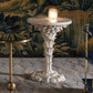 The Bunny Williams Palm Drinks Table, featuring a white sculpted base and cream stone top with candle holder, stands beside a gold-rimmed table against a patterned wall and carpeted floor.