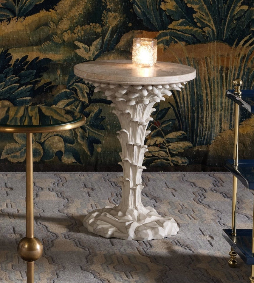 The Bunny Williams Palm Drinks Table, featuring a white sculpted base and cream stone top with candle holder, stands beside a gold-rimmed table against a patterned wall and carpeted floor.