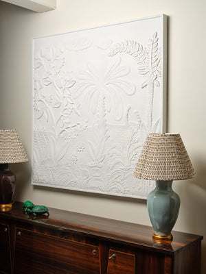 The Bunny Williams Tropical Garden Plaster Panel hangs above a wooden sideboard, flanked by two ceramic table lamps with woven beige lampshades.
