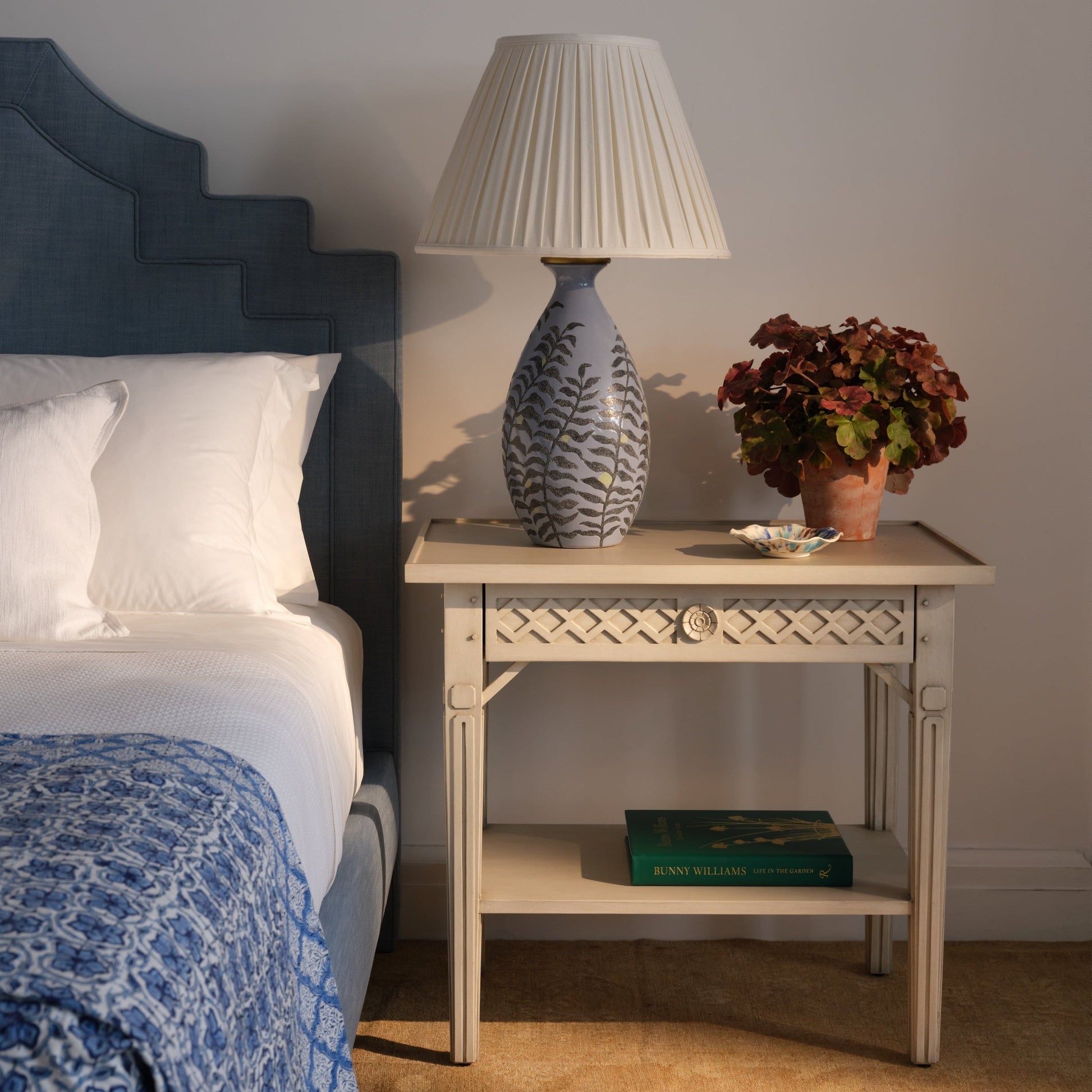 The Inge Side Table by Bunny Williams, with a hand-painted Swedish finish, displays a decorative lamp, potted plant, small dish, and green book next to a blue-upholstered bed with white bedding.