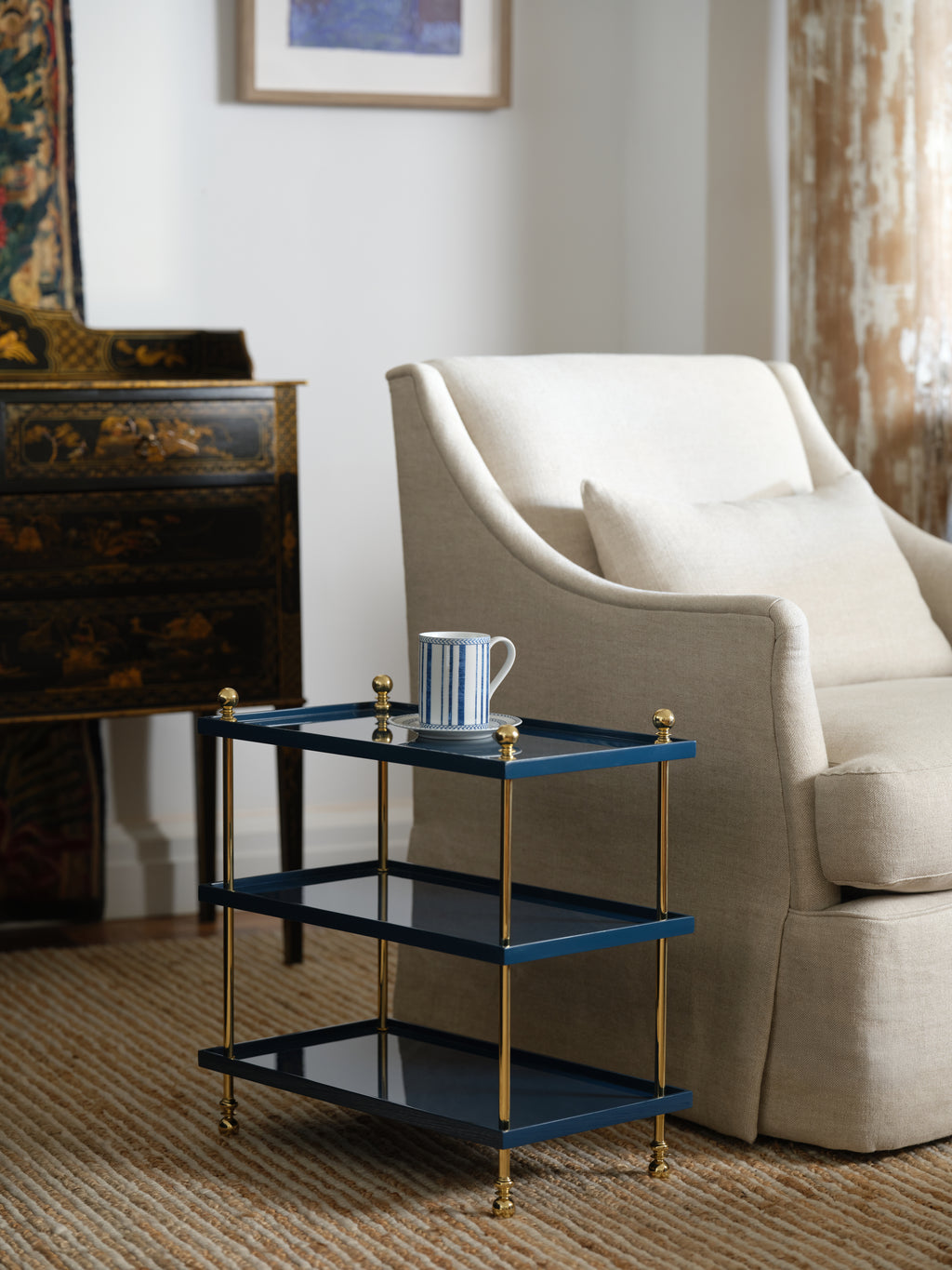 The Bunny Williams Tiered Drinks Table, finished in blue and gold lacquer with brass finials and a striped mug on top, sits beside a cream-colored armchair in the living room—an instant conversation starter.