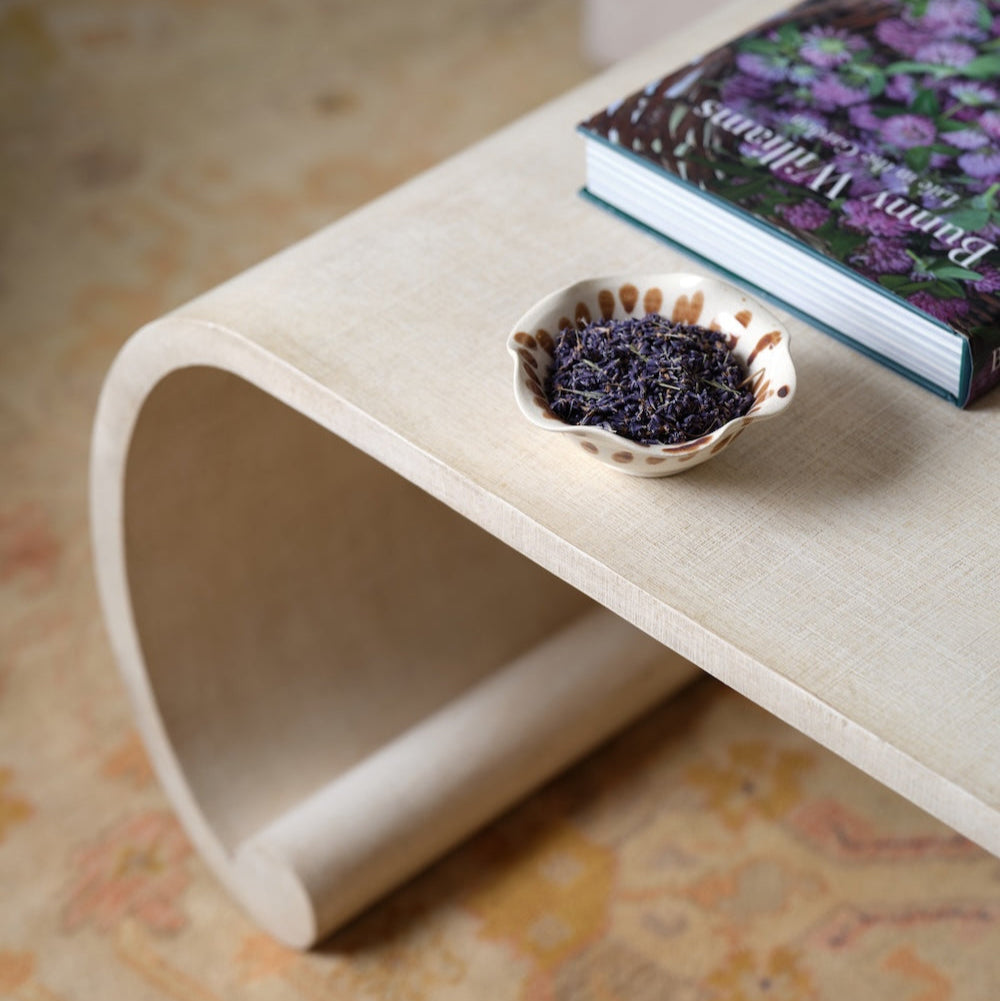 A curved beige Emilia Coffee Table by Bunny Williams with a painted linen finish, topped with a small ceramic bowl of dried lavender and a floral-covered book.
