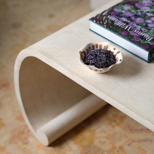 A curved beige Emilia Coffee Table by Bunny Williams with a painted linen finish, topped with a small ceramic bowl of dried lavender and a floral-covered book.