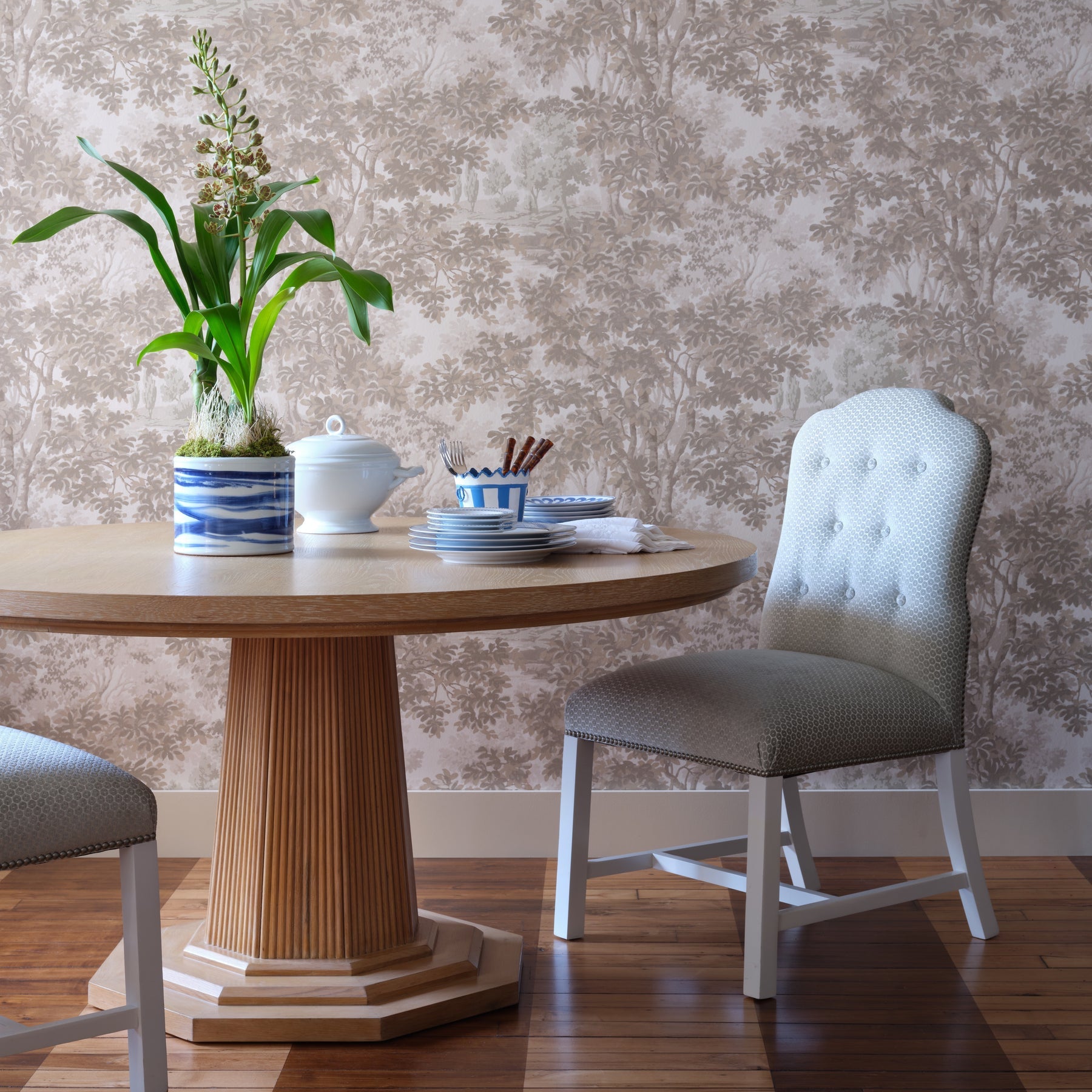 The Haven Dining Table by Bunny Williams features a pedestal base and cerused oak finish, accented with a potted plant, stacked dishes, and utensils. It’s surrounded by upholstered chairs on wood flooring with patterned wallpaper in the background.