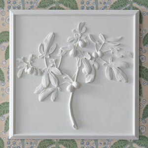 The Bunny Williams Fig Plaster Panel features a white 3D relief of a leafy fig branch with fruit, centered in a square frame and displayed against patterned wallpaper.