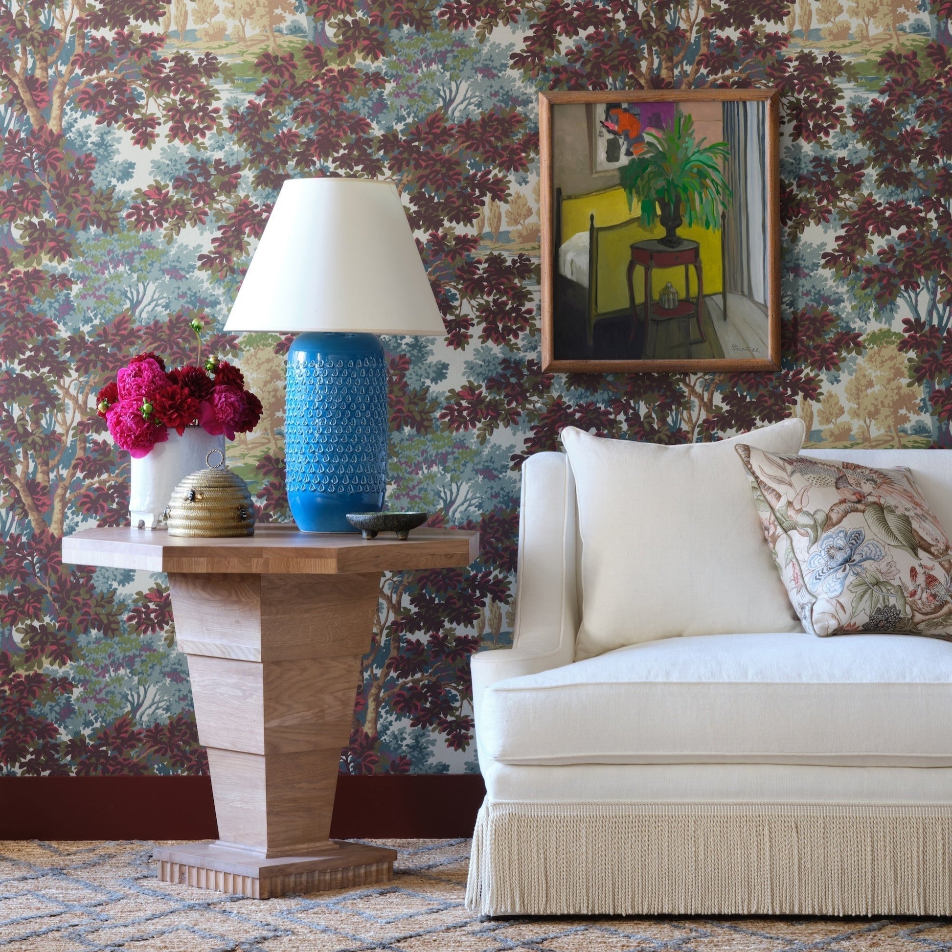 The Burton Side Table by Bunny Williams sits next to a white sofa with a patterned pillow, styled with flowers, a blue lamp, and decorative accents; floral wallpaper and framed art complete the background.