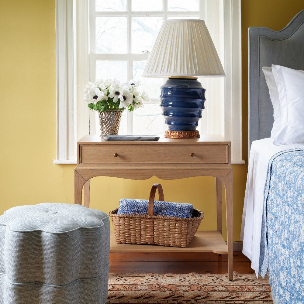 The Bunny Williams Isabella Side Table in grey-washed Oak holds a blue lamp, basket, vase of white flowers, and book beside a bed with blue bedding and a light blue upholstered ottoman.