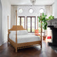 A bedroom with a Bunny Williams Rattan Trellis Canopy Bed, white bedding, fireplace, large windows, round transom windows, a potted plant, and a red upholstered chair on wood floors.