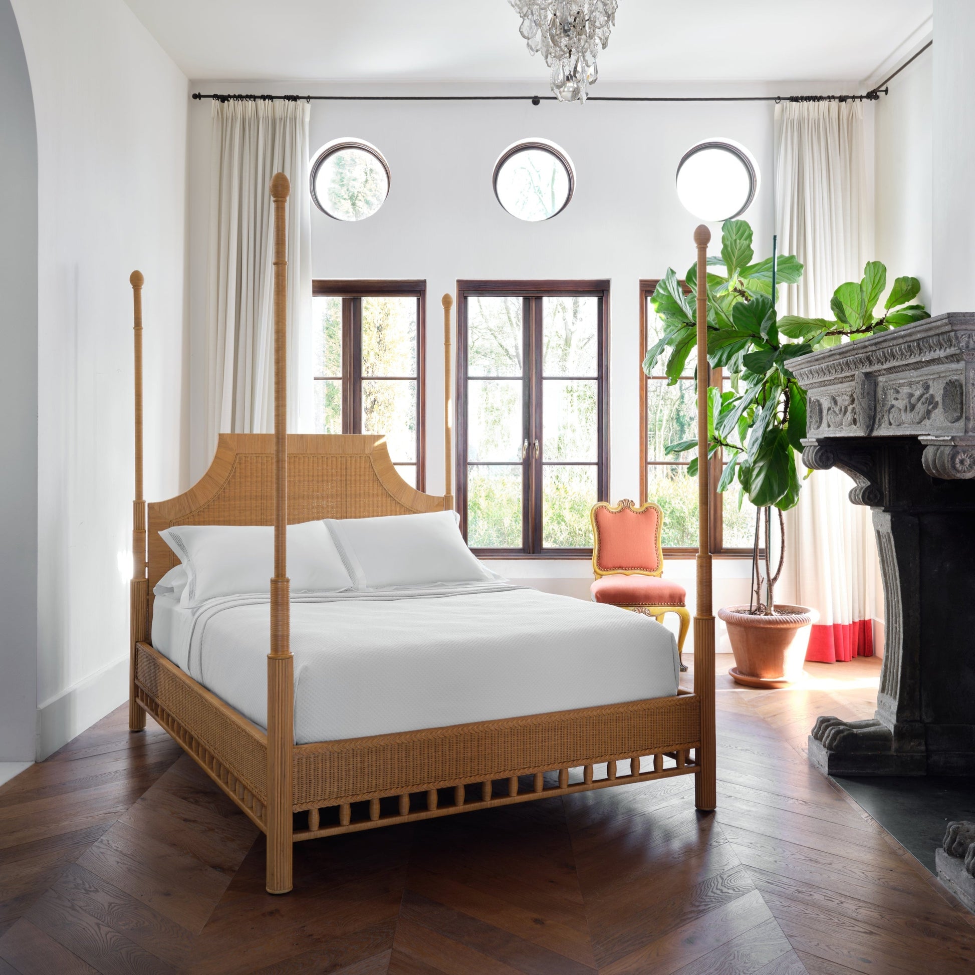 A bedroom with a Bunny Williams Rattan Trellis Canopy Bed, white bedding, fireplace, large windows, round transom windows, a potted plant, and a red upholstered chair on wood floors.