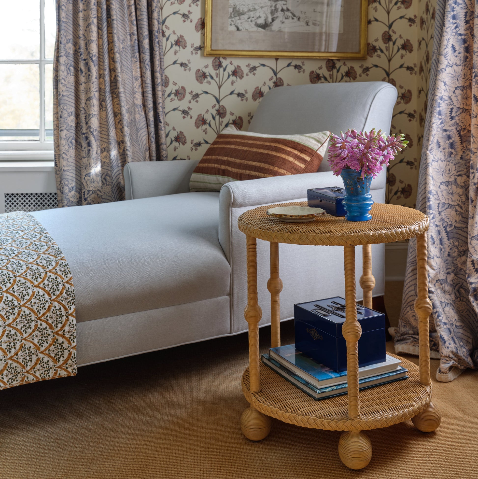 A light gray chaise lounge with a patterned throw, paired with the Bunny Williams Lucas Drinks Table—wicker, spacious, holding books and a blue vase of pink flowers—set against floral wallpaper and draped curtains.
