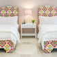 Two EJ Schrader Rosa Spring Twin XL Beds with matching colorful floral headboards and white bedding flank a white nightstand holding a lamp, books, and flowers, creating playful bedroom furniture ideal for any space.