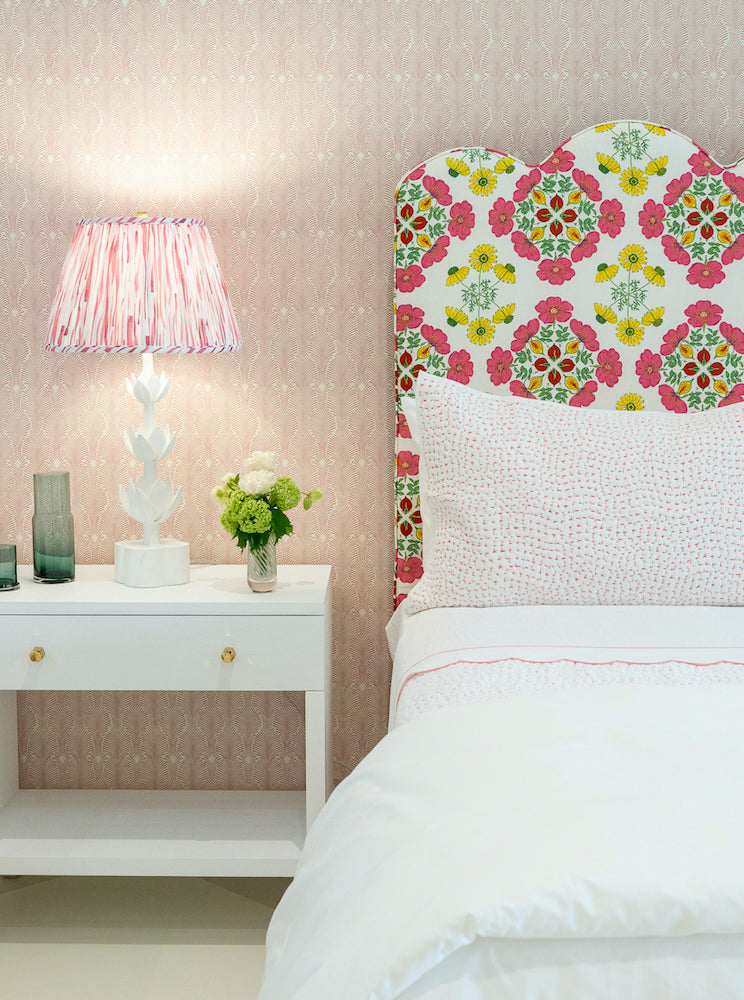 A playful bedroom features the EJ Schrader Rosa Spring Twin XL Bed with white bedding and a floral headboard, paired with a bedside table holding a lamp, glass vases, and flowers against a pink wallpapered wall.