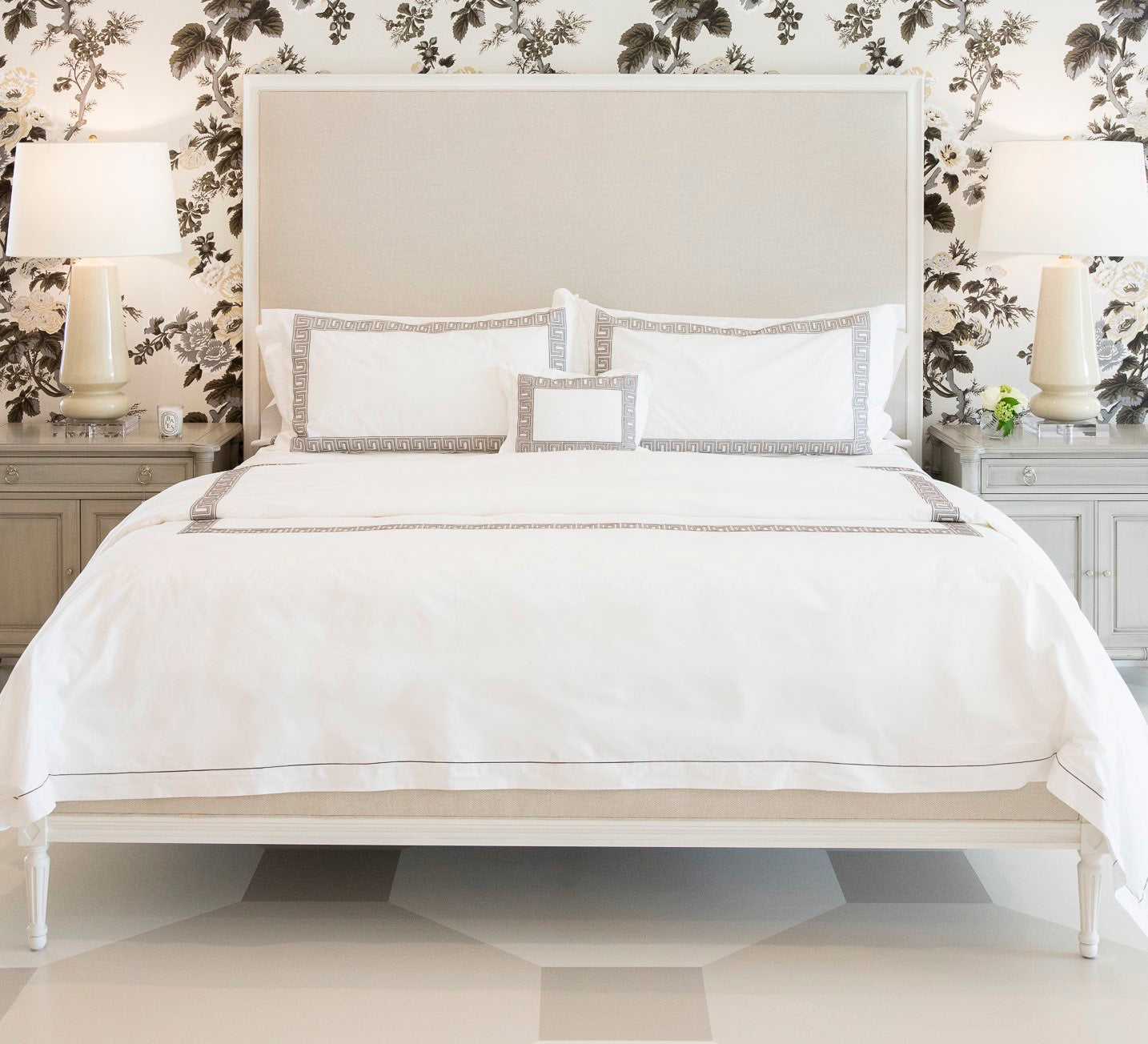 A neatly made bed features white linens with mocha trim from the Ophelia Bedding Collection by Matouk, flanked by two lamps on nightstands and set against a floral wallpaper background.