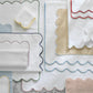Matouk’s Cairo Scallop Bath Collection, featuring bath mats and luxury long-staple cotton towels with scalloped edges in assorted colors, is artfully displayed on a marble surface.