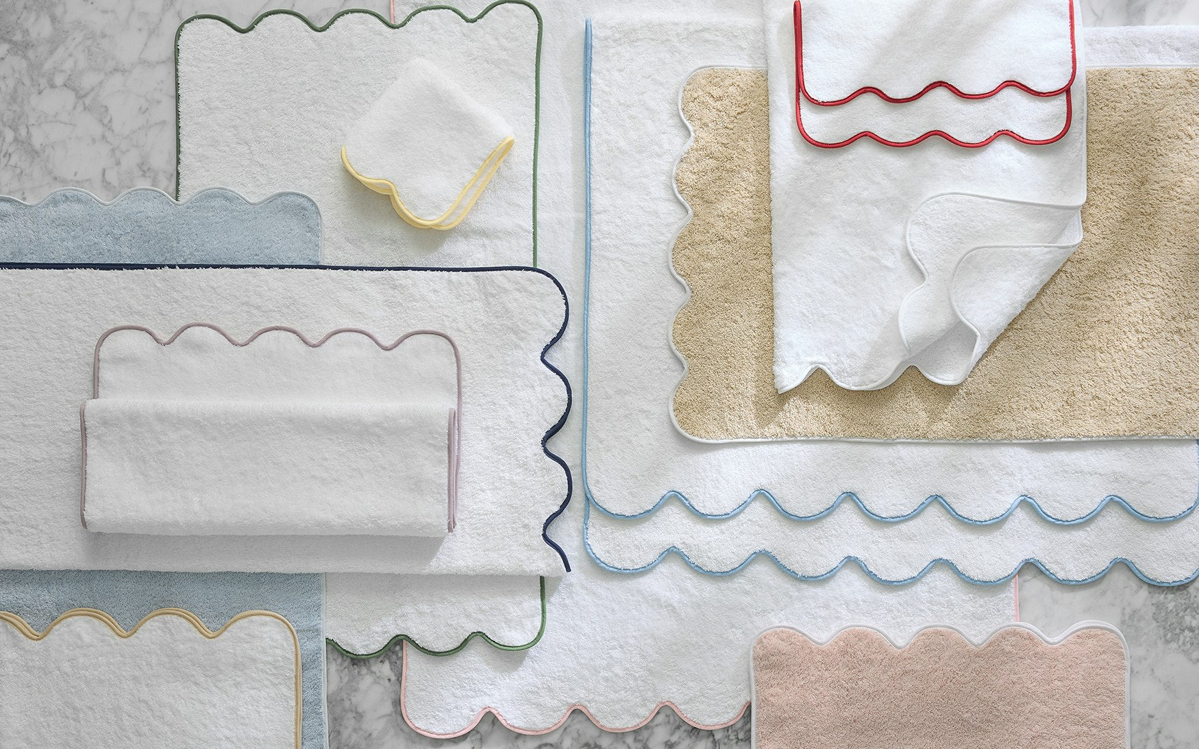 Matouk’s Cairo Scallop Bath Collection, featuring bath mats and luxury long-staple cotton towels with scalloped edges in assorted colors, is artfully displayed on a marble surface.