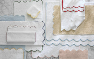 Matouk’s Cairo Scallop Bath Collection, featuring bath mats and luxury long-staple cotton towels with scalloped edges in assorted colors, is artfully displayed on a marble surface.