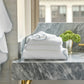A stack of Matouk's Jasper Bath Collection white towels with grey trim rests on a grey marble bathroom counter beside a gold faucet and a silver cup filled with green flowers.