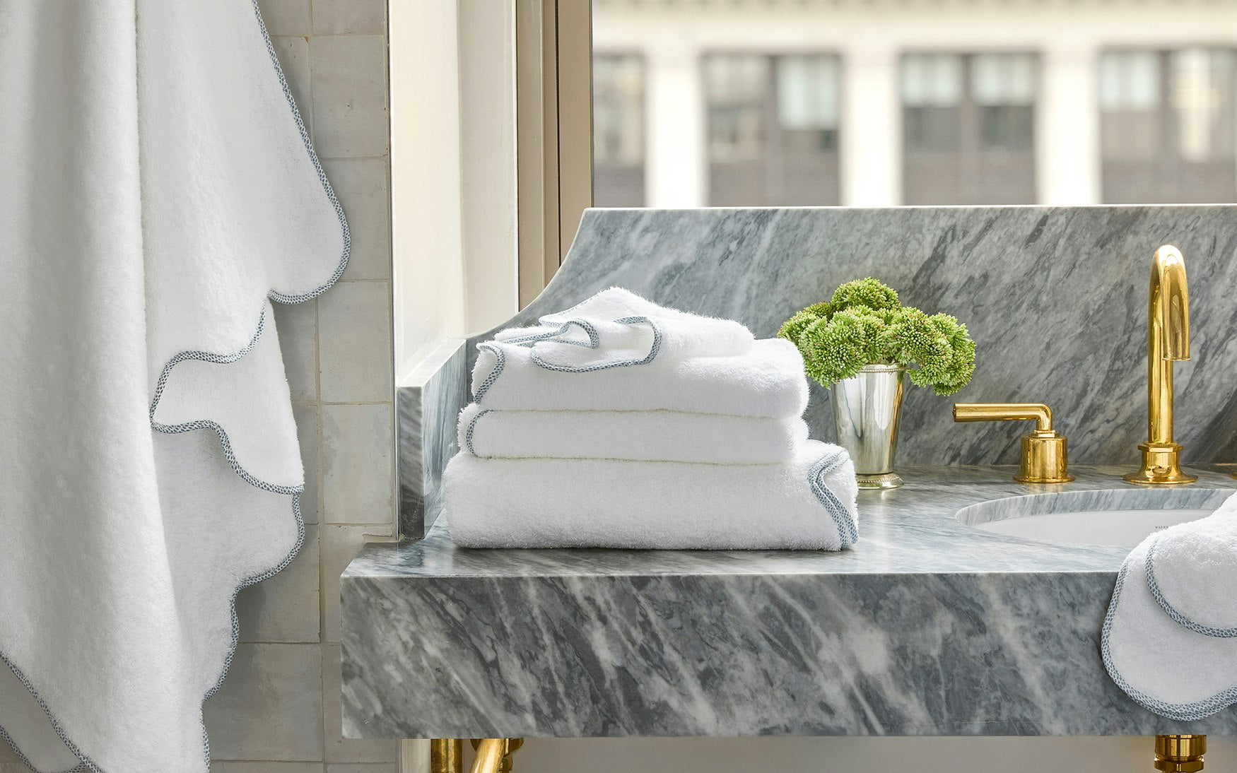 A stack of Matouk's Jasper Bath Collection white towels with grey trim rests on a grey marble bathroom counter beside a gold faucet and a silver cup filled with green flowers.