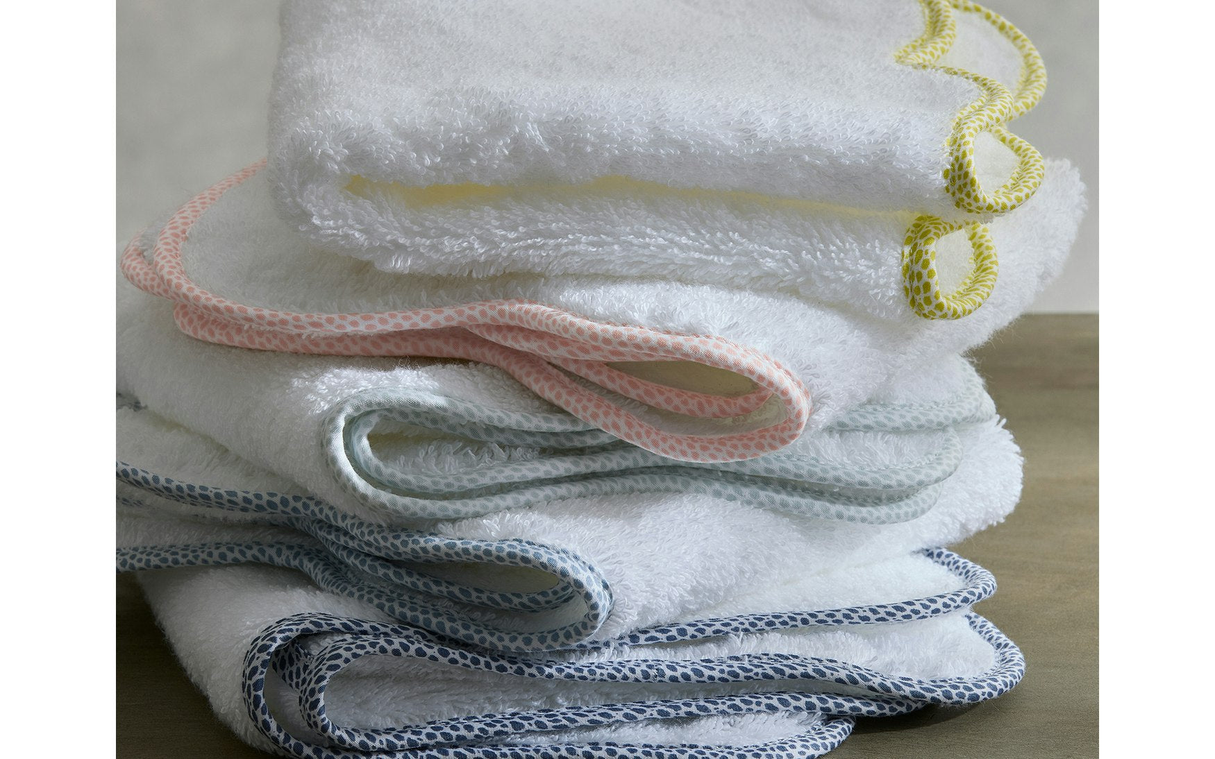 A stack of Jasper Bath Collection towels by Matouk, featuring white fabric with scalloped edges and colored trim in yellow, pink, light blue, and dark blue, is beautifully arranged on a wooden surface.