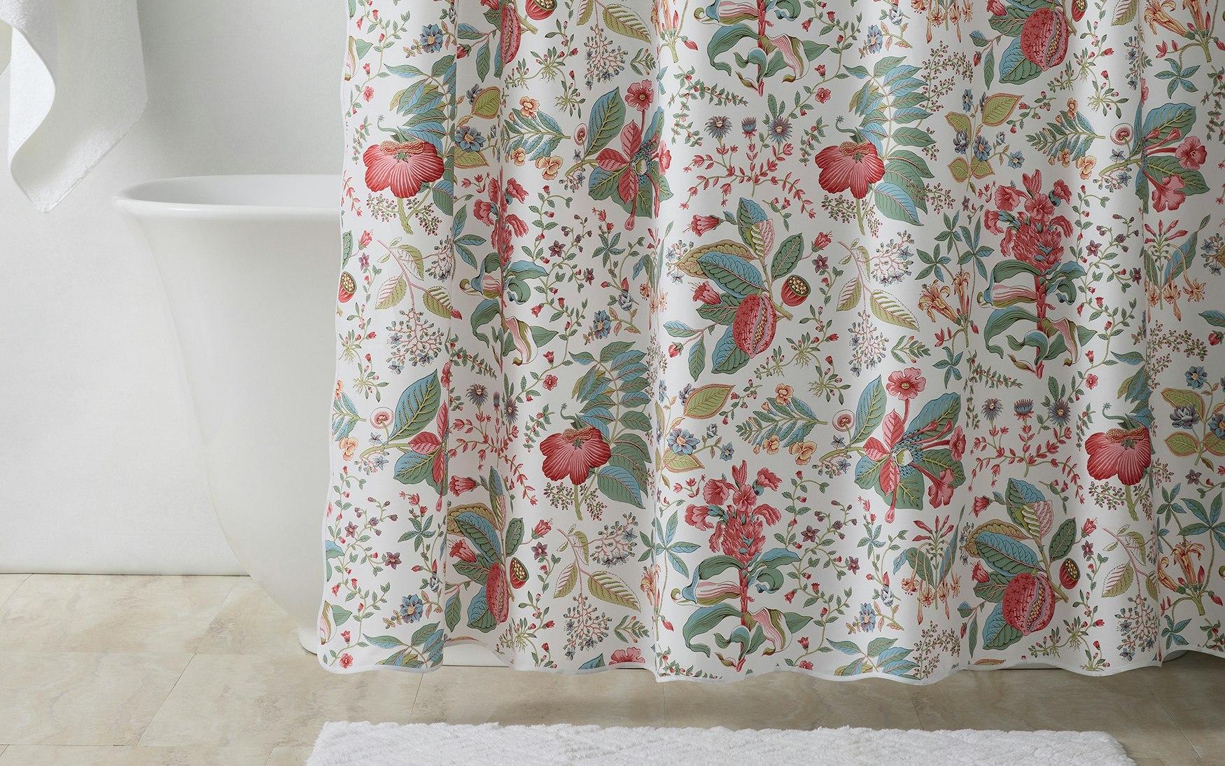 A white shower curtain from the Matouk Pomegranate Shower Curtain Collection, featuring a vibrant botanical floral pattern inspired by Schumacher designs, hangs in front of a white bathtub; a coordinating white towel and bath mat complete the look.