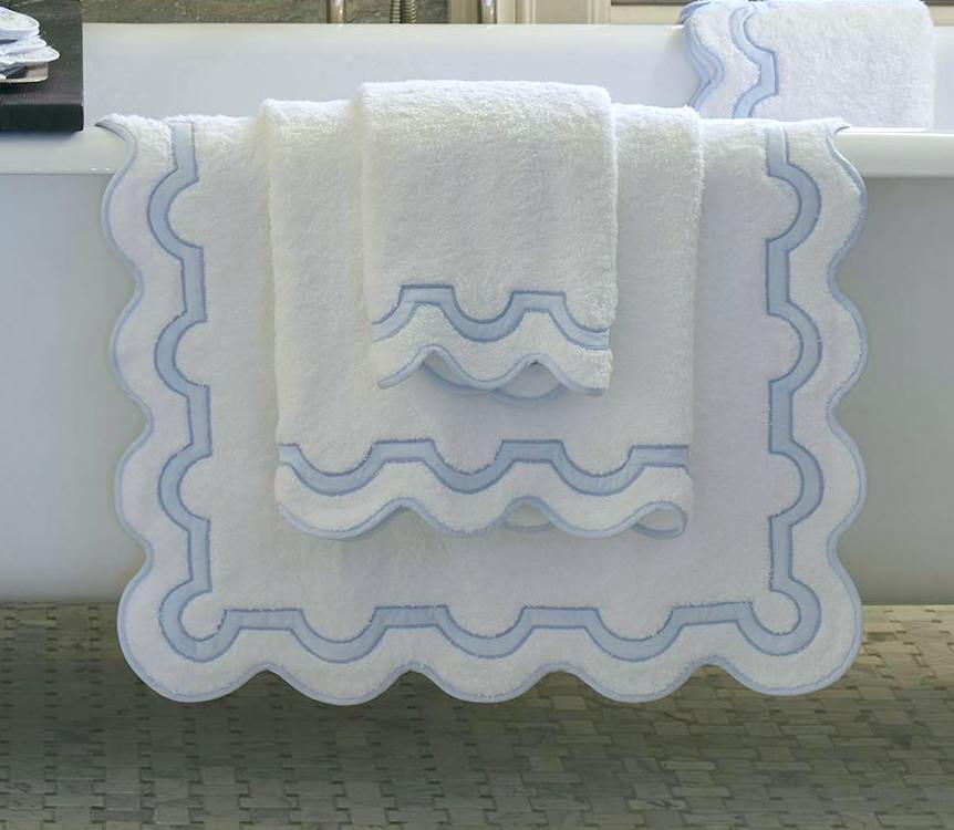 Matouk’s Mirasol Bath Collection towels, featuring scalloped blue-trimmed edges, are draped over a white bathtub, with another Cairo cotton terry towel visible on the edge in the background.