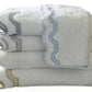 A stack of white Matouk Mirasol Bath Collection towels with scalloped edges in gray, blue, and beige trim, neatly folded against a plain background.
