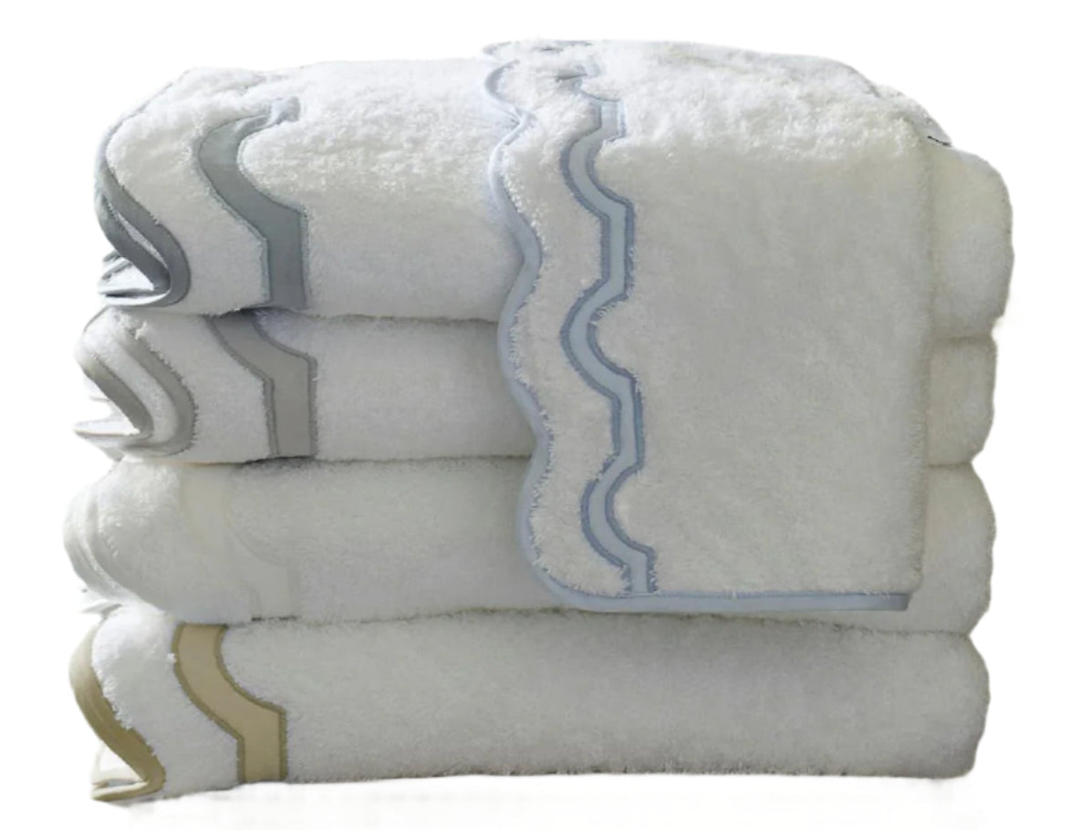 A stack of white Matouk Mirasol Bath Collection towels with scalloped edges in gray, blue, and beige trim, neatly folded against a plain background.