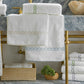 Matouk’s Classic Chain Bath Collection, featuring white cotton terry towels with signature chain embroidery, is displayed on and beneath a marble sink with gold piping, accompanied by a basket and a decorative stone bowl.