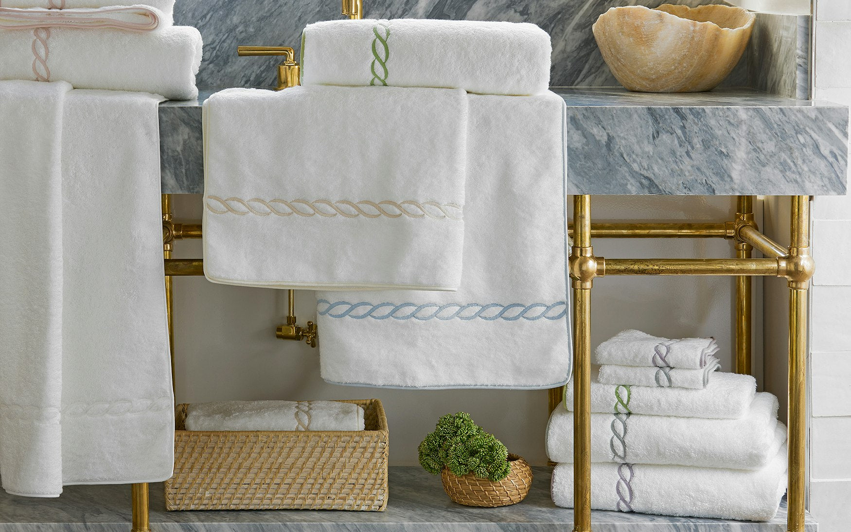 Matouk’s Classic Chain Bath Collection, featuring white cotton terry towels with signature chain embroidery, is displayed on and beneath a marble sink with gold piping, accompanied by a basket and a decorative stone bowl.