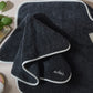 The Giza Makeup Towel Set by Matouk features fade-resistant dark gray towels with white trim labeled “makeup,” displayed on a light wood surface beside a body brush, green leaves, and cosmetics.
