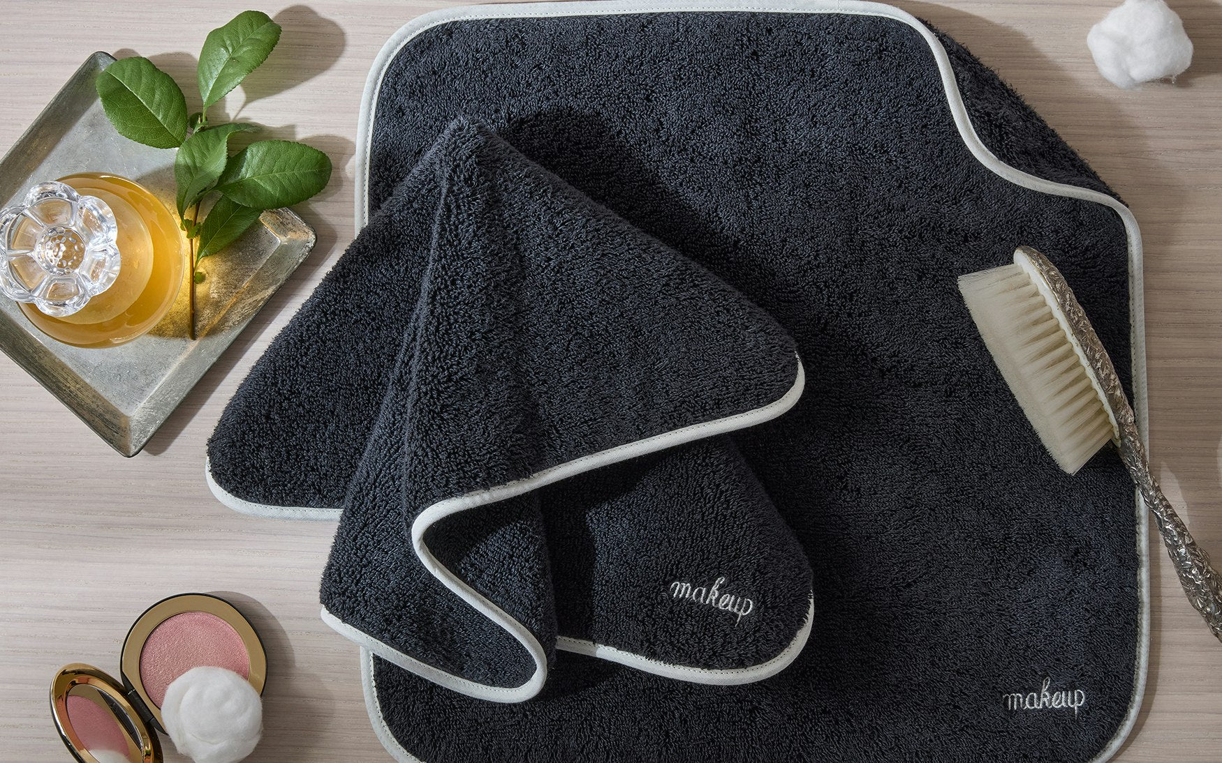 The Giza Makeup Towel Set by Matouk features fade-resistant dark gray towels with white trim labeled “makeup,” displayed on a light wood surface beside a body brush, green leaves, and cosmetics.