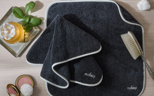 The Giza Makeup Towel Set by Matouk features fade-resistant dark gray towels with white trim labeled “makeup,” displayed on a light wood surface beside a body brush, green leaves, and cosmetics.
