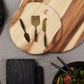 The Be Home Arendal Cheese Knife Set features three ethically crafted knives with black handles, displayed on a round, sustainably sourced wooden board beside another board, a gray placemat, and a black salad bowl.