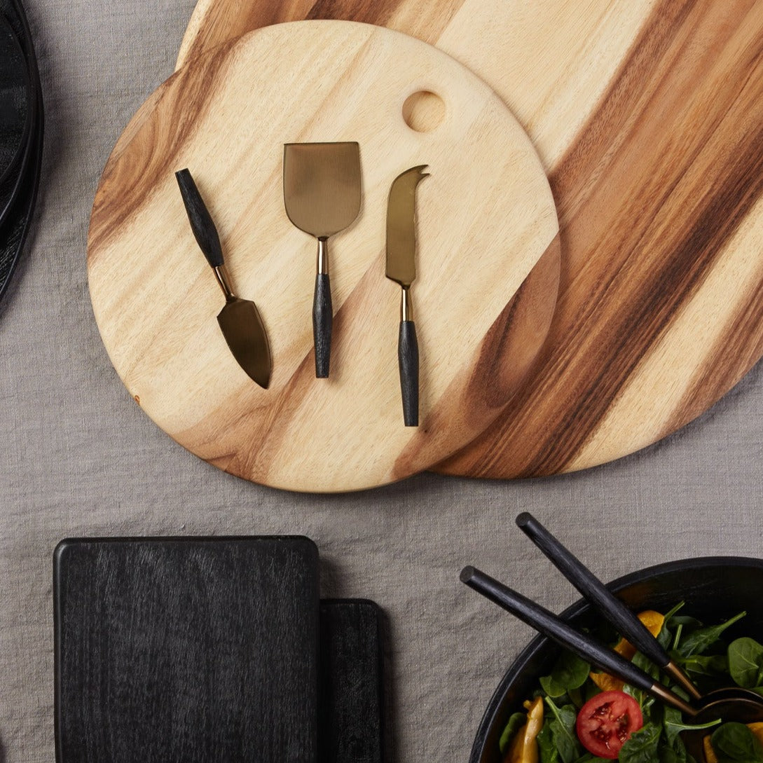 The Be Home Arendal Cheese Knife Set features three ethically crafted knives with black handles, displayed on a round, sustainably sourced wooden board beside another board, a gray placemat, and a black salad bowl.