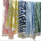 Five towels from the Matouk Zebra Beach Towel Collection in pastel pink, yellow, navy blue, light blue, and green with white tropical leaf patterns hang side by side on a line.